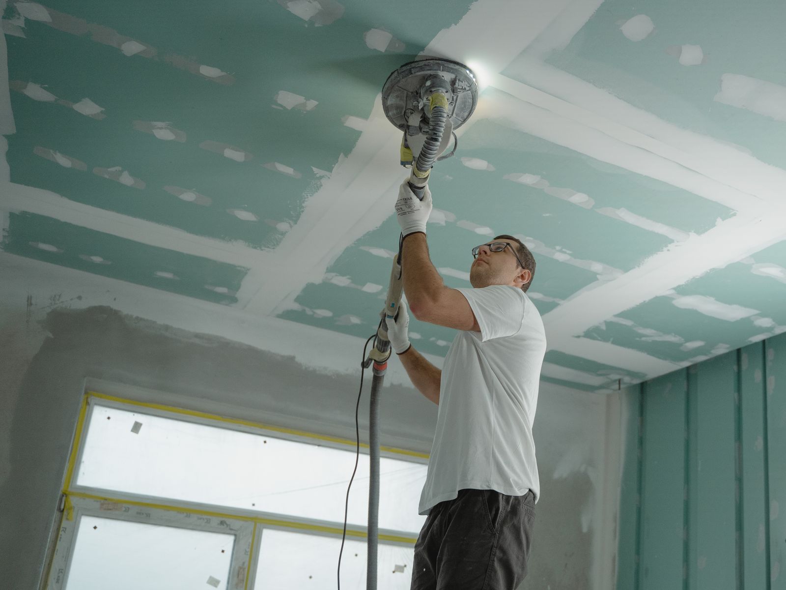Contractor using a dust-extracting sander to smooth a skim-coated drywall ceiling during refinishing